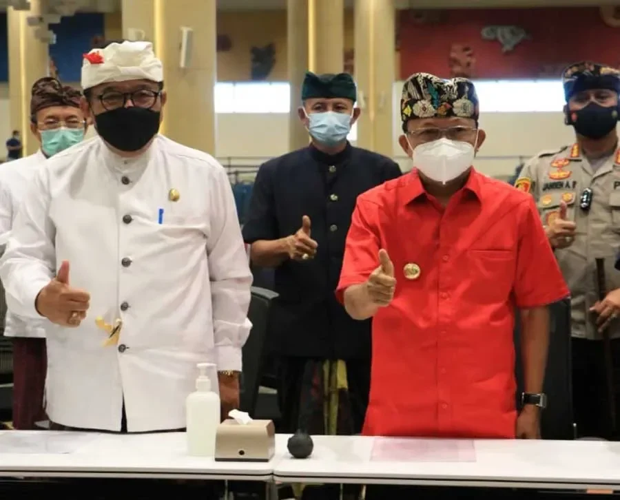 Dua pria memberikan jempol, memakai pakaian adat dan masker di dalam ruangan.