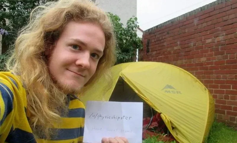 Seorang pria berambut pirang tersenyum di depan tenda kuning dengan tulisan Reddit.